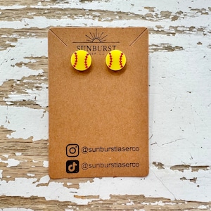May include: A pair of yellow softball stud earrings with red stitching, displayed on a brown card. The card has the text "SUNBURST LASER CO" and social media handles. The earrings are round and resemble miniature softballs.