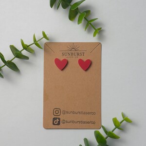 May include: A pair of red heart-shaped stud earrings on a brown card with the text "SUNBURST LASER CO" and social media handles for @sunburstlaserco.