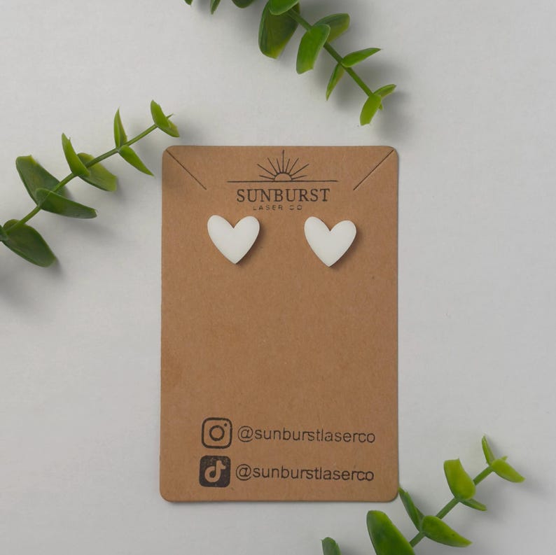 May include: White heart-shaped stud earrings on a brown card with the text "SUNBURST LASER CO" and social media handles for @sunburstlaserco.