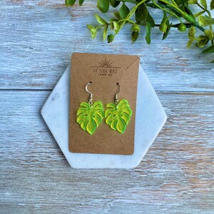 May include: A pair of green and yellow monstera leaf earrings with silver hooks. The earrings are displayed on a brown card with the text "Sunburst Laser Co."
