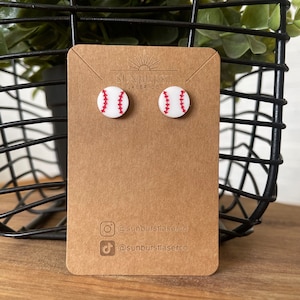 May include: A pair of white baseball earrings with red stitching. The earrings are displayed on a brown card with the text "Sunburst Laser Co." and social media handles.