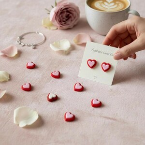 May include: Red and pink heart-shaped earrings on a white card held by a hand. Scattered around are small heart-shaped candies, a bracelet, rose petals, and a cup of coffee. The card reads "Sunburst Laser Co."