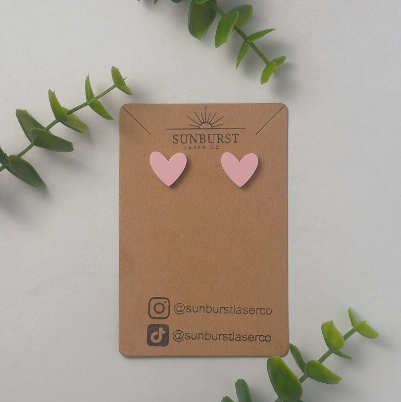 May include: A pair of pink heart-shaped earrings on a brown card with the text "SUNBURST LASER CO" and social media handles for @sunburstlaserco.
