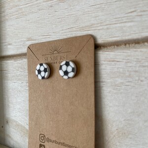 May include: A pair of white and black soccer ball stud earrings on a brown card with the text "Sunburst Laser Co." and "@sunburstlaserco".