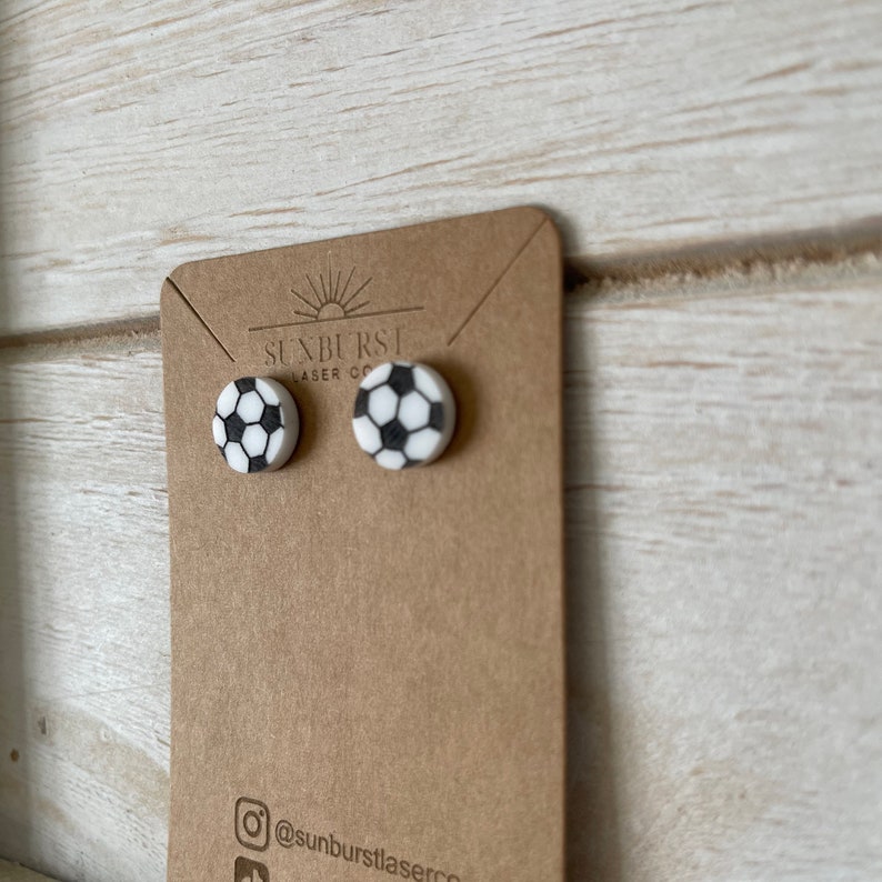 May include: A pair of white and black soccer ball stud earrings on a brown card with the text "Sunburst Laser Co." and "@sunburstlaserco".