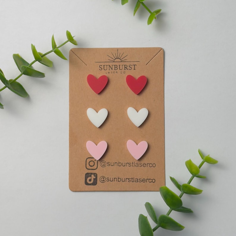 May include: A set of six heart-shaped earrings in red, white, and pink. The earrings are displayed on a brown card with the text "Sunburst Laser Co." and social media handles.