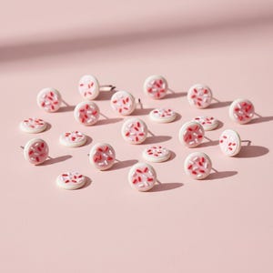 May include: A collection of small, round stud earrings and flat disc earrings. The earrings are white with a pink center and red and white sprinkles. The background is a solid light pink.