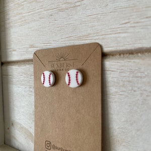 May include: A pair of white baseball earrings with red stitching. The earrings are on a brown card with the text "Sunburst Laser Co." and a sun graphic.