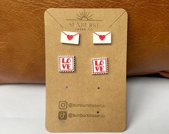 May include: A set of four wooden earrings featuring a red heart on a white envelope and a red "LOVE" stamp on a white square. The earrings are displayed on a brown card with the text "SUNBURST LASER CO" and social media handles.