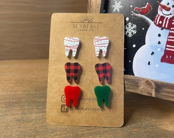 May include: A set of six Christmas-themed tooth earrings. Two pairs are made of wood with a red and white striped pattern and a red and black plaid pattern. The other two earrings are made of acrylic and are red and green.