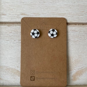 May include: A pair of white and black soccer ball stud earrings on a brown card with the text "Sunburst Laser Co." and social media handles for Instagram and TikTok.