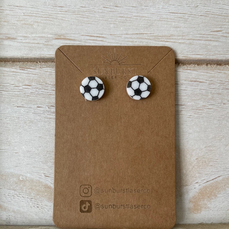 May include: A pair of white and black soccer ball stud earrings on a brown card with the text "Sunburst Laser Co." and social media handles for Instagram and TikTok.