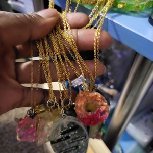 Op de afbeelding: Een verzameling goudkleurige kettingkettingen met verschillende hangerontwerpen. De hangers bevatten heldere, roze en zwarte harsstukken, sommige met glitter en tekst. De kettingen worden in een hand gehouden, met een blauw rek op de achtergrond.