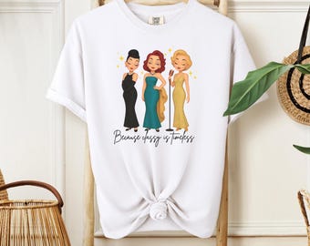 Può includere: T-shirt bianco con una grafica di tre donne in abiti da sera, ognuna con un microfono. Il testo "Because classy is timeless" è scritto sotto. La maglietta è appesa a una gruccia di legno.