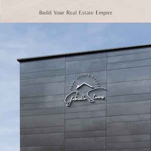 May include: A silver real estate agent logo on a gray building. The logo features a house icon, the text "Real Estate Agent", and the name "Gabrielle Stevens" in cursive.