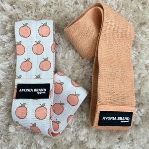 May include: Two resistance bands. One is white with a peach pattern and the other is solid peach. Both bands have a black label with the text "AVONIA BRAND equipment". The bands are laying on a white, fluffy surface.