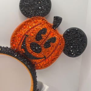 May include: Black and orange Mickey Mouse ears with a pumpkin design covered in rhinestones. The pumpkin has a smiling face with black eyes and a wide grin.