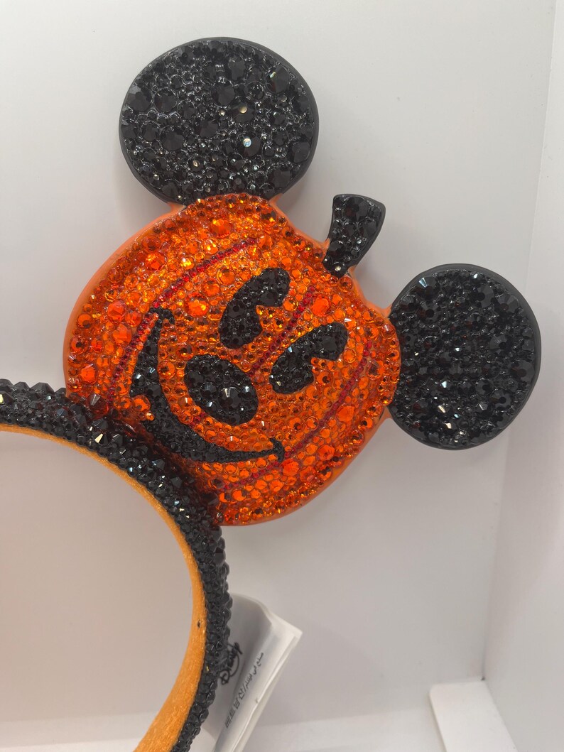 May include: Black and orange Mickey Mouse ears with a pumpkin design covered in rhinestones. The pumpkin has a smiling face with black eyes and a wide grin.