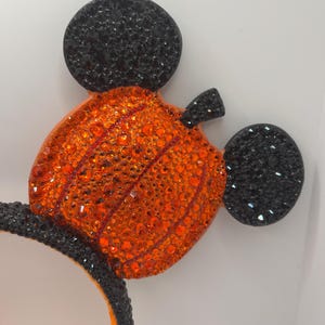 May include: A pair of black and orange Mickey Mouse ears with a pumpkin design. The ears are covered in rhinestones.