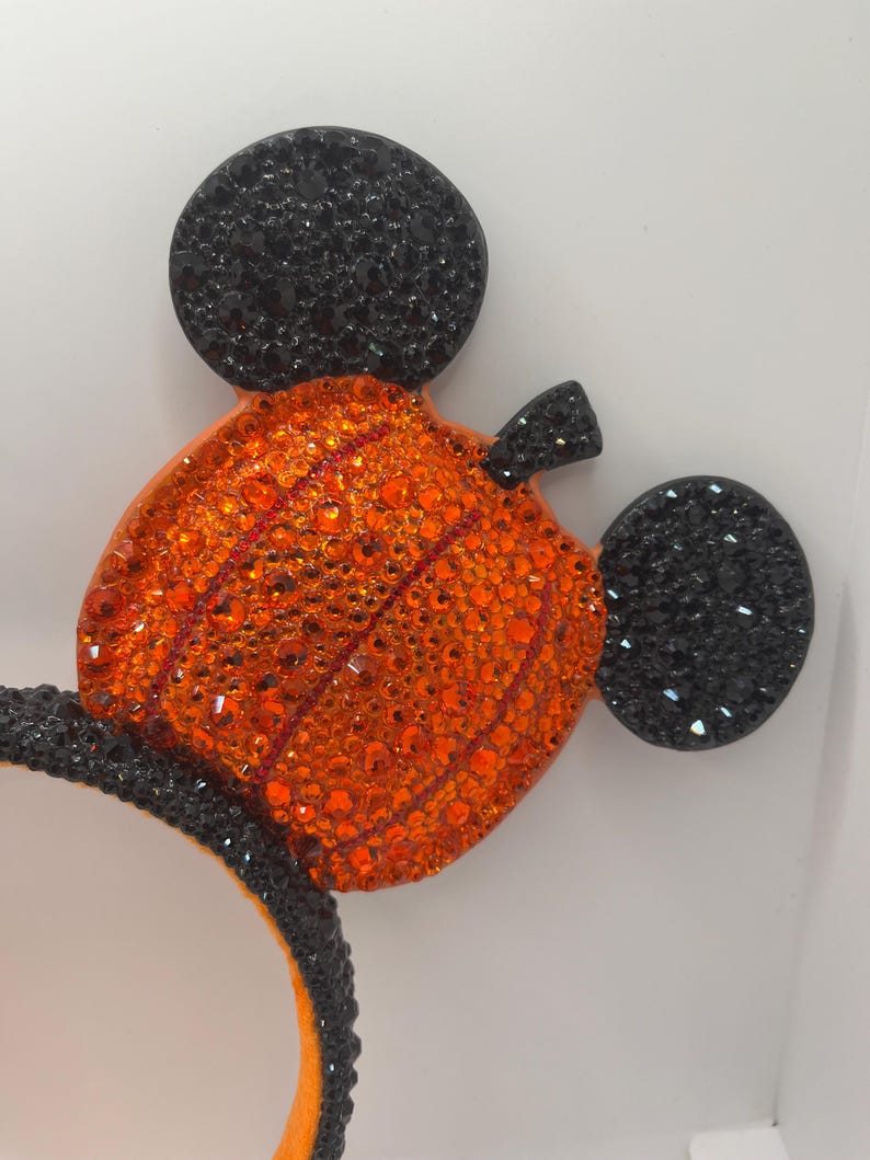 May include: A pair of black and orange Mickey Mouse ears with a pumpkin design. The ears are covered in rhinestones.