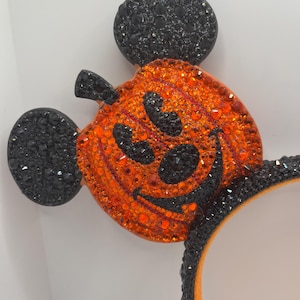 May include: Black and orange sparkly Mickey Mouse ears with a pumpkin design. The pumpkin has a black face with a wide smile and eyes.