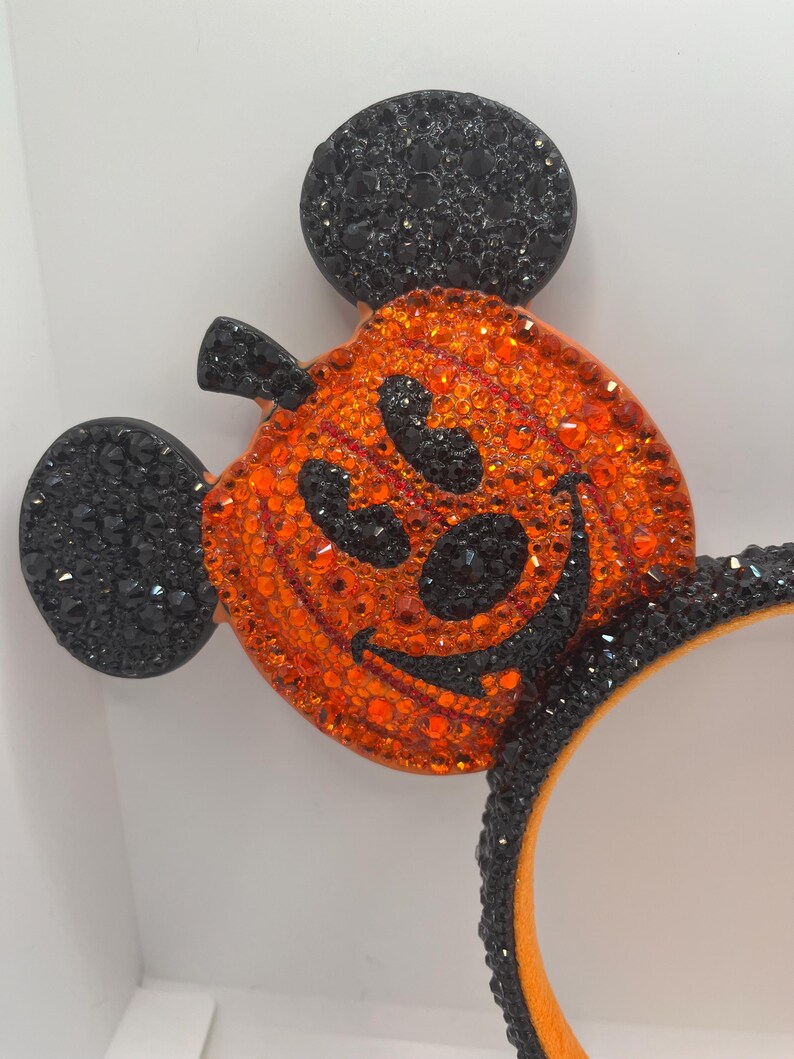 May include: Black and orange sparkly Mickey Mouse ears with a pumpkin design. The pumpkin has a black face with a wide smile and eyes.