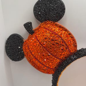 May include: Black and orange Mickey Mouse ears with a pumpkin design made of rhinestones. The pumpkin is orange and has a green stem. The ears are covered in black rhinestones.