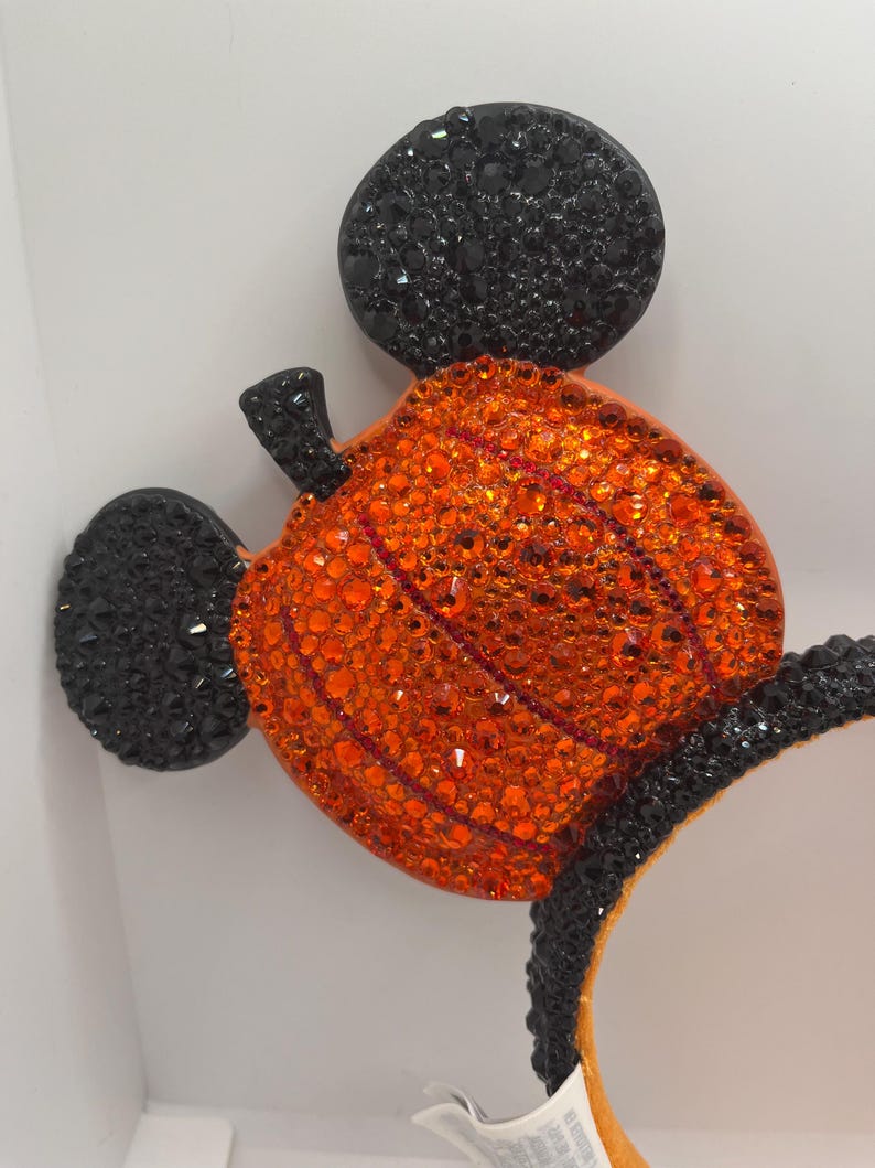 May include: Black and orange Mickey Mouse ears with a pumpkin design made of rhinestones. The pumpkin is orange and has a green stem. The ears are covered in black rhinestones.