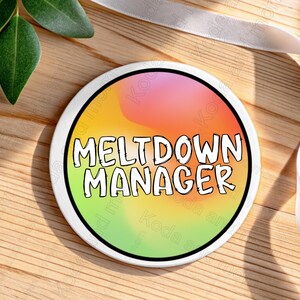 Könnte beinhalten: Ein runder, weißer Untersetzer mit dem Text "MELTDOWN MANAGER" in fetten, schwarzen Buchstaben. Der Untersetzer hat einen farbenfrohen Hintergrund mit Rosa-, Gelb- und Grüntönen. Er liegt auf einer Holzoberfläche.