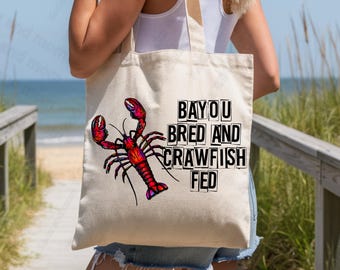 May include: A beige tote bag featuring a vibrant red and purple crawfish illustration. The text "BAYOU BRED AND CRAWFISH FED" is printed in black block letters. The bag is carried outdoors near a beach.