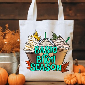 May include: White tote bag with a graphic of three coffee drinks with whipped cream and the text "BASIC BITCH SEASON" in teal. The bag is surrounded by pumpkins and fall foliage.