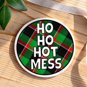 以下が含まれることがあります： 緑、黒、赤のチェック柄の丸いコースター。「HO HO HOT MESS」の文字が白い太字で縦に重ねられています。白い縁取りがあり、お祝いの季節に最適です。
