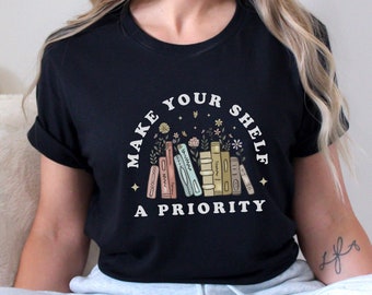 May include: Black t-shirt with a white graphic that says "Make your shelf a priority". The graphic features a stack of colorful books with flowers and stars.