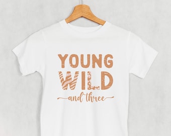May include: White t-shirt with the text "YOUNG WILD and three" in a brown, distressed font. The word "WILD" has a patterned design. The shirt is hanging on a wooden hanger against a light gray background.