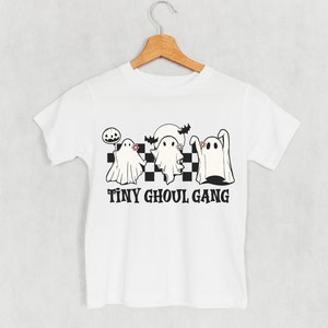 Peut inclure: T-shirt blanc avec un motif sur le thème d'Halloween. Le motif présente trois fantômes de dessins animés, une citrouille et le texte "TINY GHOUL GANG" en noir. Les fantômes sont sur un motif à carreaux noir et blanc.