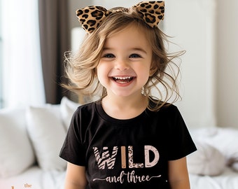 May include: A young girl wearing a black t-shirt with the text "WILD and three" in pink and gold letters. The letters are decorated with animal print patterns. She is wearing a leopard print headband and smiling.