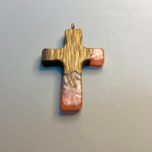 May include: A wooden cross pendant with a pink and white resin inlay. The cross is a religious symbol and is often used as a piece of jewelry.