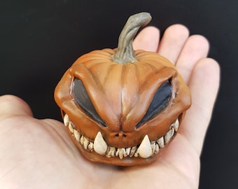 May include: A small, orange ceramic pumpkin with a menacing grin and black eyes. The pumpkin has a detailed, sculpted face with sharp teeth.