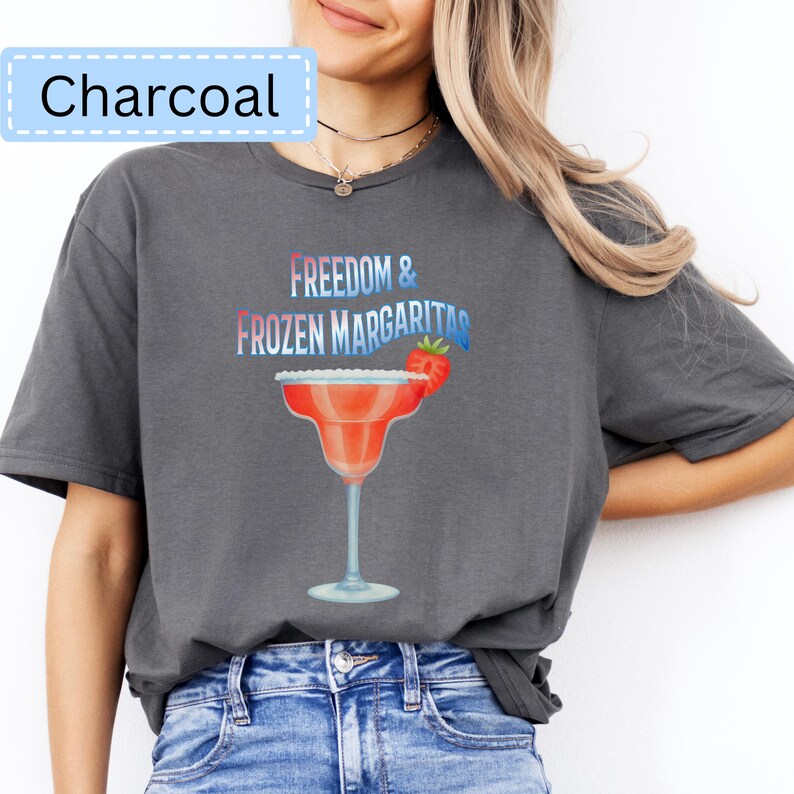 May include: Charcoal-colored t-shirt featuring a margarita glass graphic with the words "Freedom & Frozen Margaritas." The graphic includes a red margarita glass with a strawberry garnish. The shirt is a comfortable, casual style.