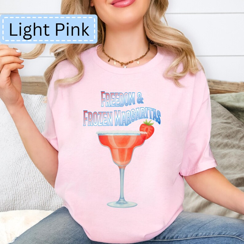 May include: Light pink t-shirt with the text "Freedom & Frozen Margaritas" in blue and red. A watercolor illustration of a margarita glass with a strawberry is featured on the front.