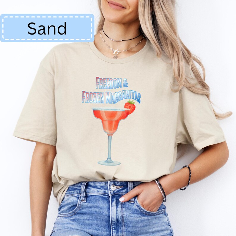 May include: Sand-colored t-shirt featuring the text "Freedom & Frozen Margaritas" above a margarita glass with a strawberry garnish. The shirt has a relaxed fit and short sleeves. The text "Sand" is in a light blue box.