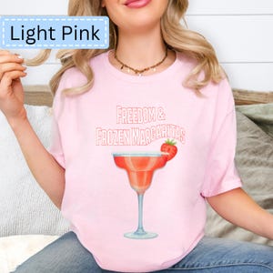 May include: Light pink t-shirt with the text "Freedom & Frozen Margaritas" and a watercolor illustration of a margarita with a strawberry garnish. The shirt is light pink.