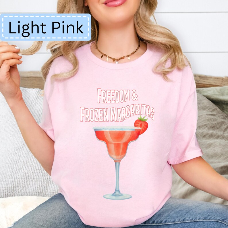 May include: Light pink t-shirt with the text "Freedom & Frozen Margaritas" and a watercolor illustration of a margarita with a strawberry garnish. The shirt is light pink.