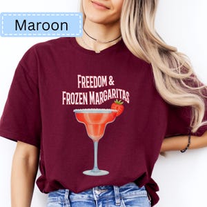 May include: Maroon t-shirt with the text "Freedom & Frozen Margaritas" and a margarita glass graphic. The margarita glass is red with a strawberry garnish. The shirt is a solid maroon color.