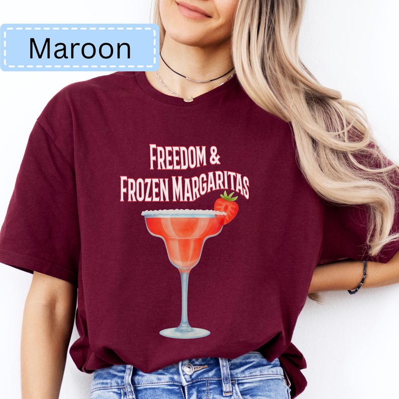 May include: Maroon t-shirt with the text "Freedom & Frozen Margaritas" and a margarita glass graphic. The margarita glass is red with a strawberry garnish. The shirt is a solid maroon color.