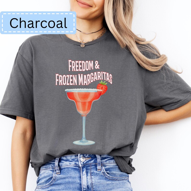 May include: Charcoal-colored t-shirt with the text "Freedom & Frozen Margaritas" in pink. A red margarita glass with a strawberry garnish is printed on the front. The shirt is a casual, comfortable style.