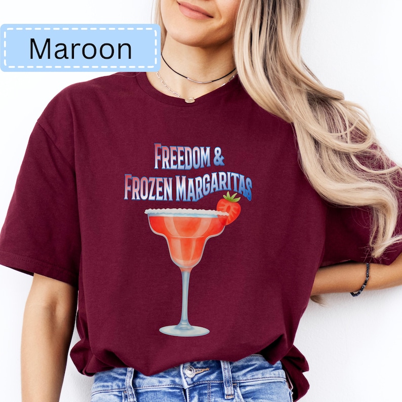 May include: Maroon t-shirt with the text "Freedom & Frozen Margaritas" and a margarita glass graphic. The margarita glass is red with a white rim and a strawberry garnish. The shirt is a solid maroon color.