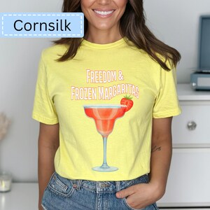 May include: A yellow t-shirt with the text "Freedom & Frozen Margaritas" and a graphic of a margarita glass with a strawberry. The shirt also has the word "Cornsilk" in a light blue box.