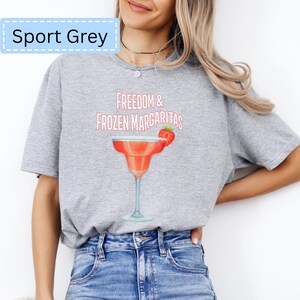 May include: Sport Grey t-shirt with the text "Freedom & Frozen Margaritas" and a watercolor illustration of a margarita cocktail with a strawberry garnish. The shirt is a light gray color.