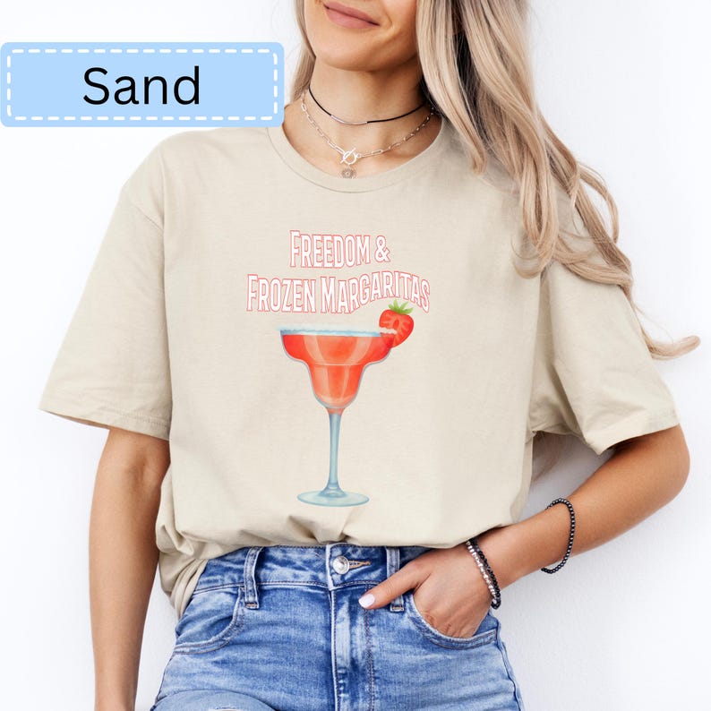 May include: Sand-colored t-shirt featuring the text "Freedom & Frozen Margaritas" above a margarita glass with a strawberry garnish. The shirt has a relaxed fit and short sleeves. The word "Sand" is in a light blue box in the upper left corner.